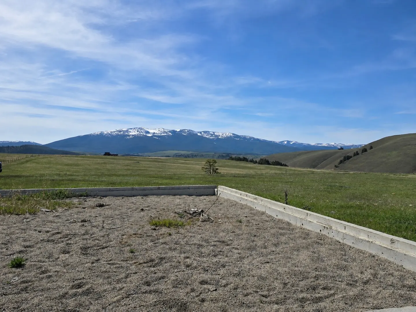 A foundation on a grassy lot with snow-capped mountains in the background under a blue sky.