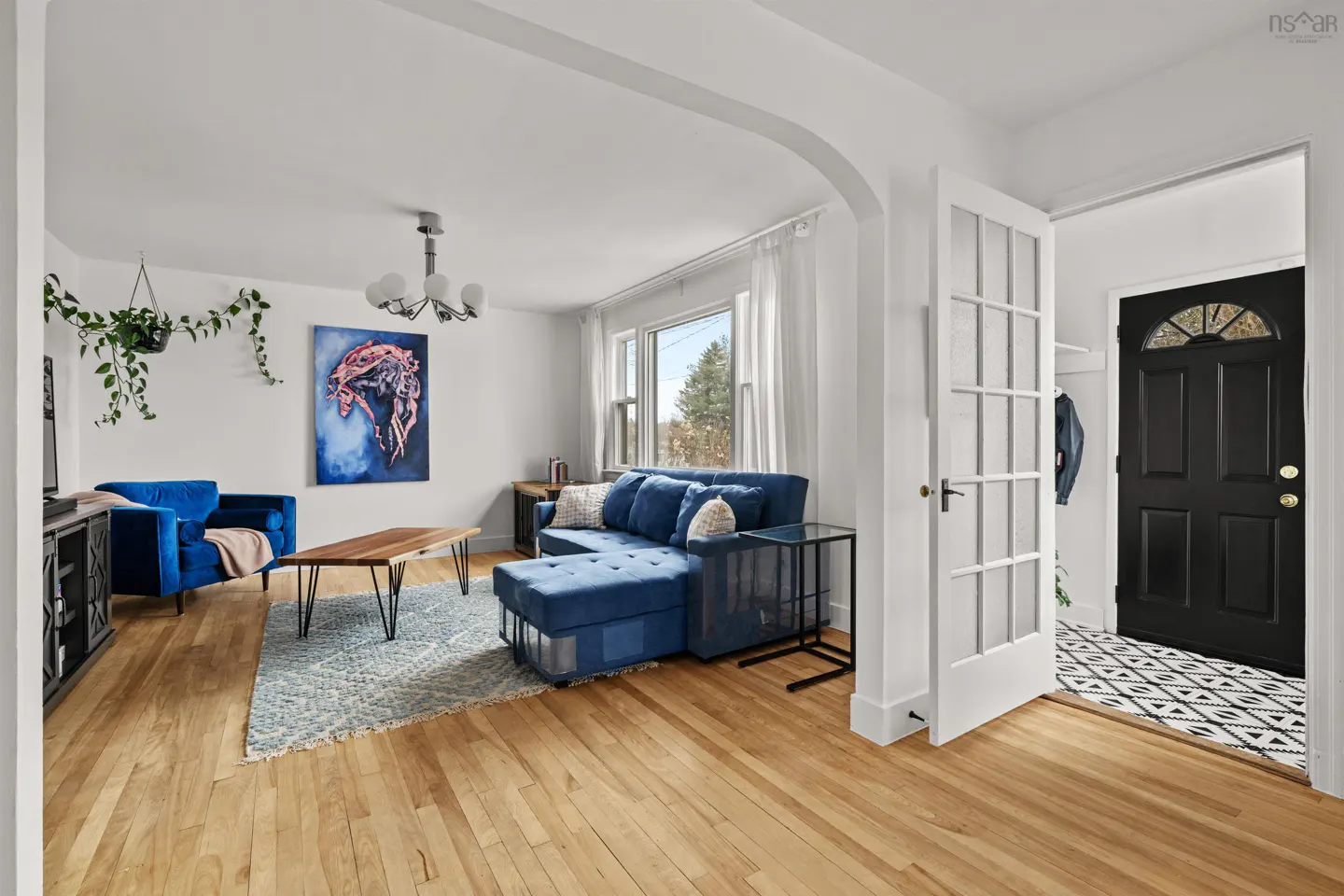 Bright living room with hardwood floors, blue velvet sofa and chair, and a black front door with patterned rug.