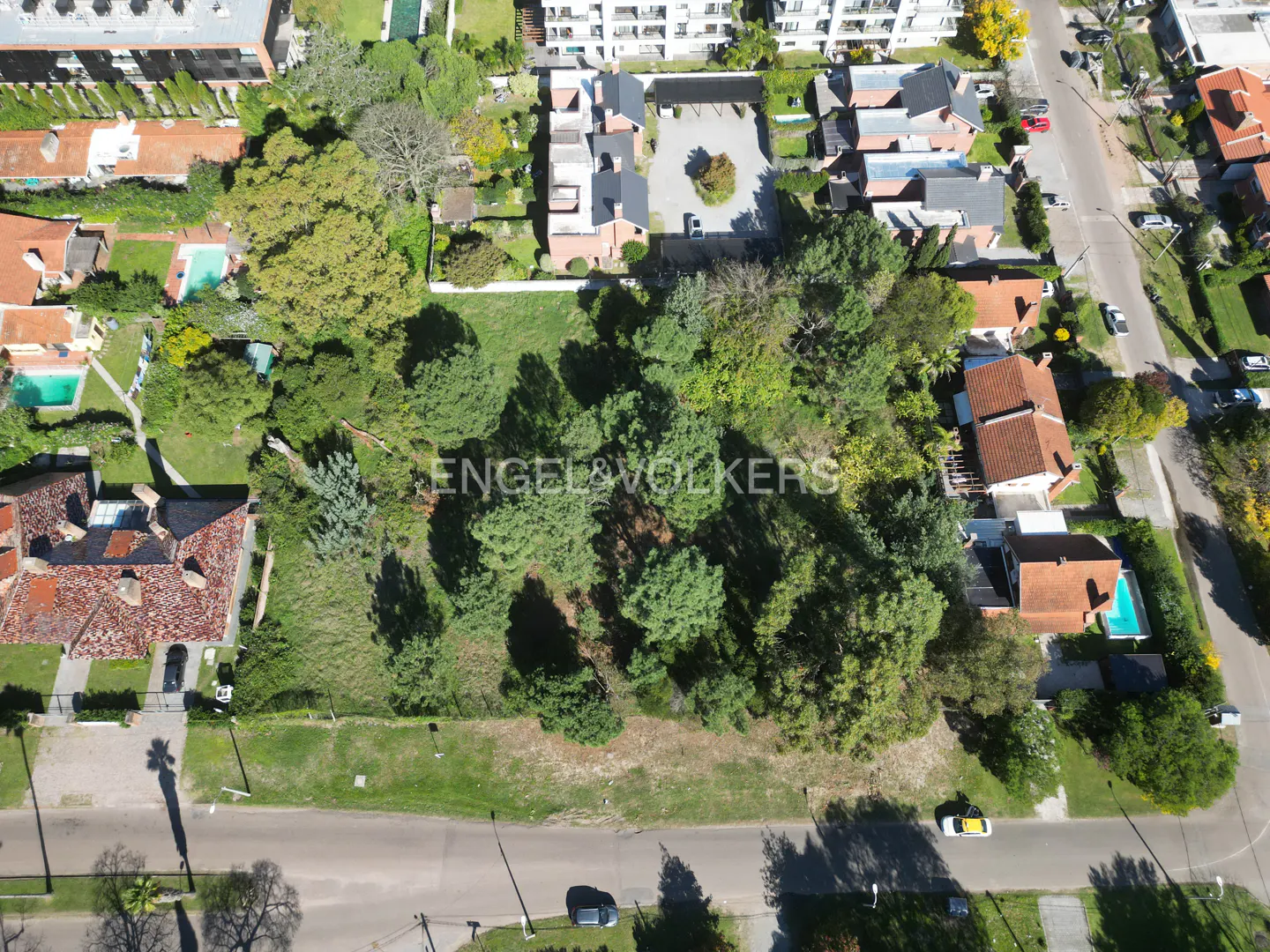 Aerial view of a green, grassy lot with trees, surrounded by houses and streets in a residential area. Engel & Völkers logo is visible.