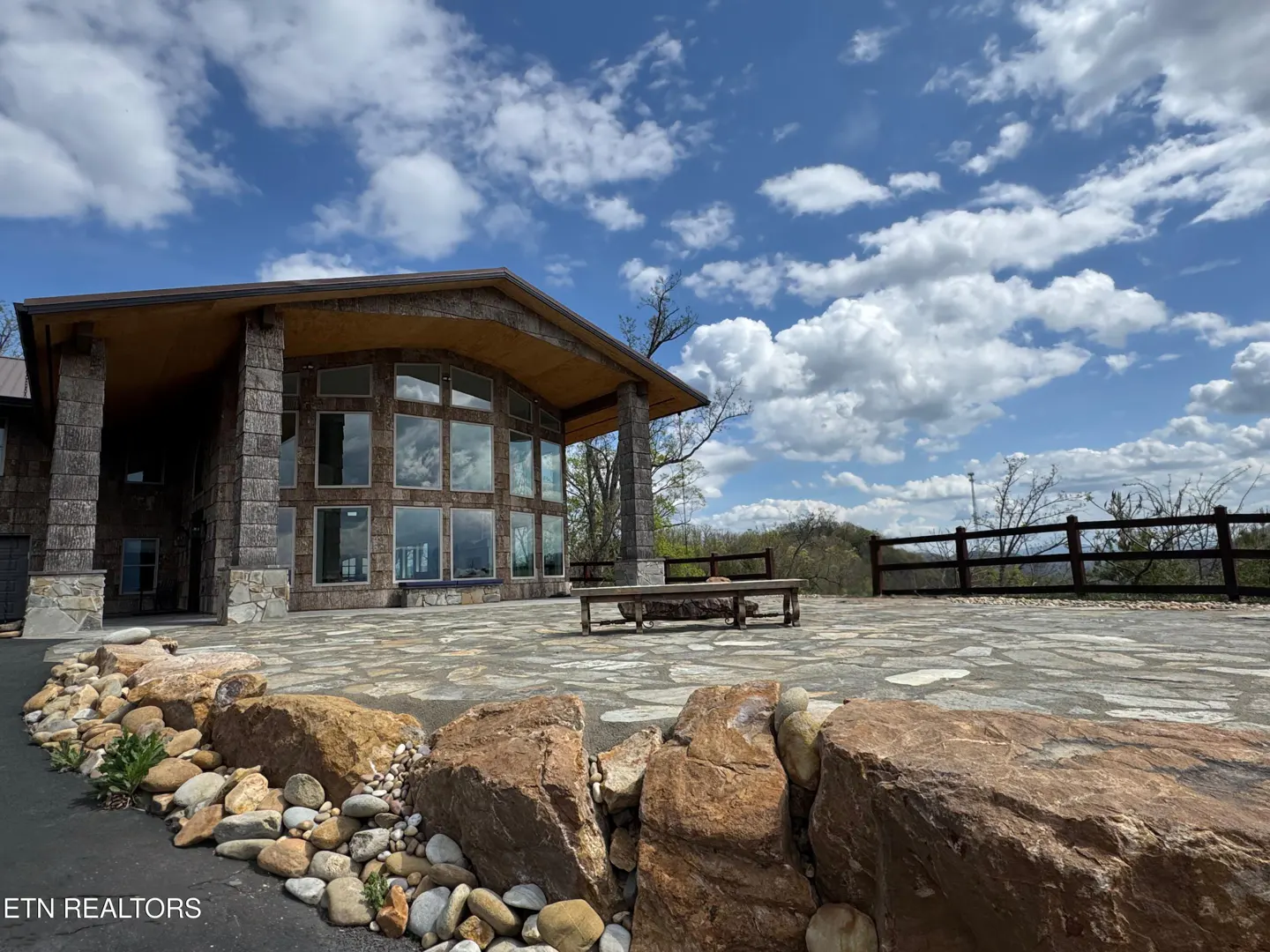 Stone house with large windows and patio. A wooden bench sits on the stone patio overlooking a green landscape under a blue sky with clouds.