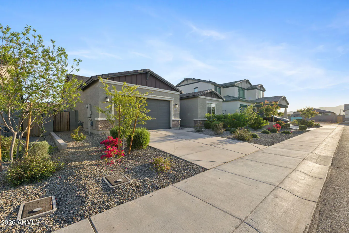 A modern, single-story home with a gray exterior, dark roof, and a two-car garage. Landscaping includes gravel, bushes, and small trees. Blue sky above.