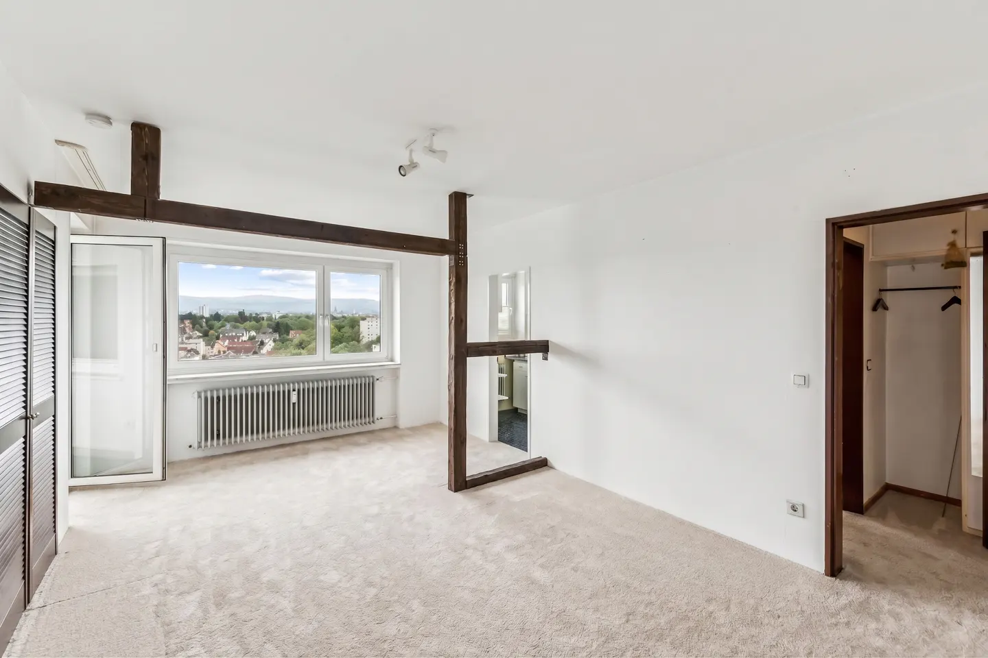 Bright, empty room with white walls, beige carpet, and a large window with a city view. Dark wood beams accent the space.