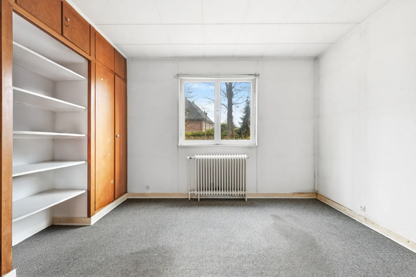 Empty room with gray carpet, white walls, and a window. A wooden closet with shelves is on the left. A radiator is under the window.