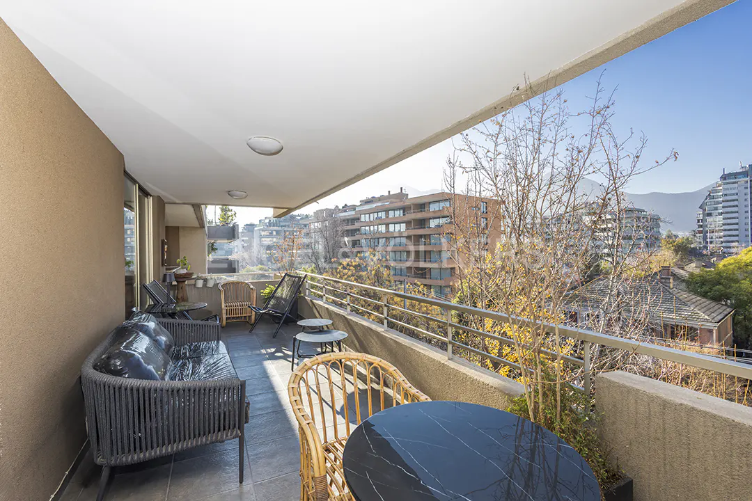 A spacious balcony with outdoor furniture, including a sofa, chairs, and a table, overlooks a cityscape with buildings and trees.