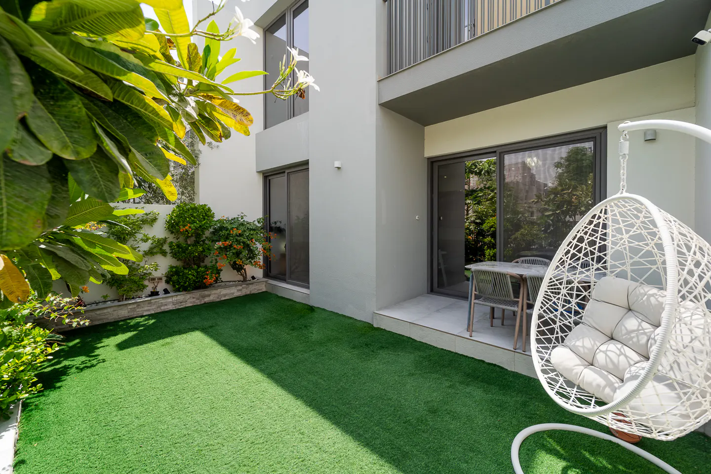 A modern home's backyard with green grass, a white hanging chair, and a patio with a table and chairs.