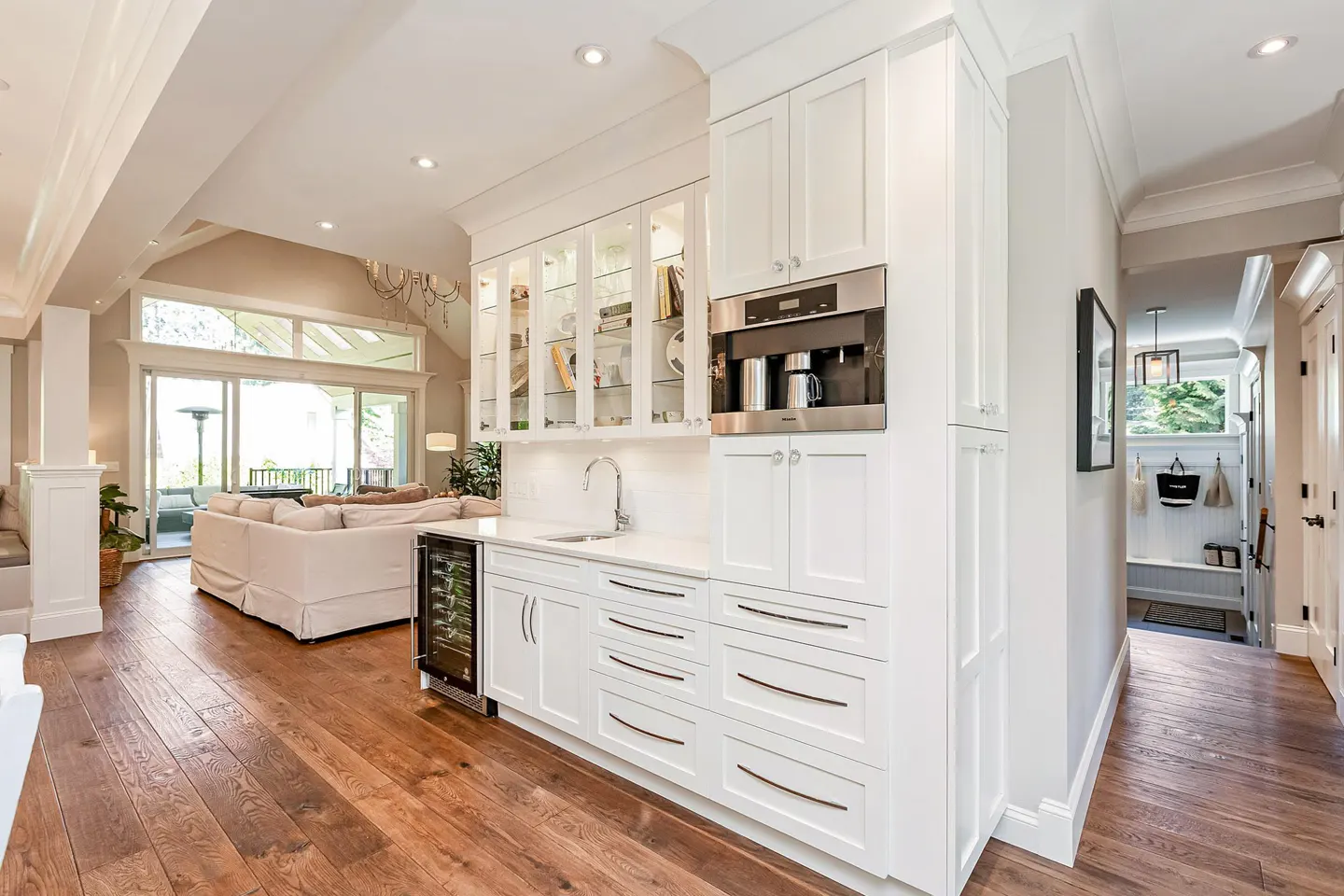 A bright, modern home interior with white cabinets, a built-in coffee maker, and a wine fridge. A living room and hallway are visible.