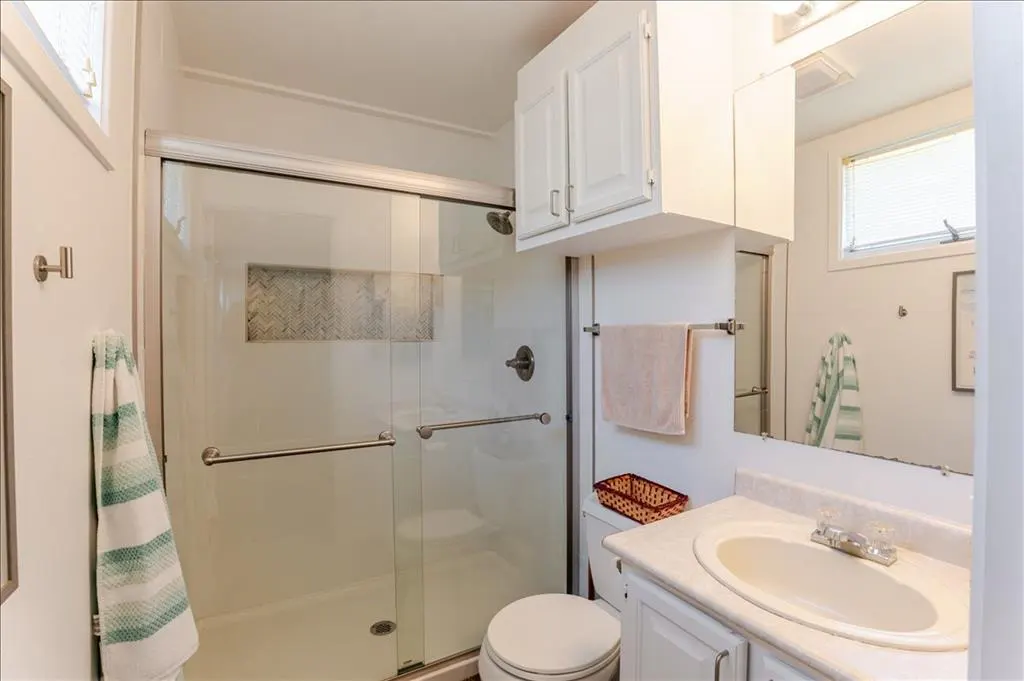 Bright bathroom with white cabinets, sink, and toilet. Glass shower with silver hardware and a towel rack with a pink towel.
