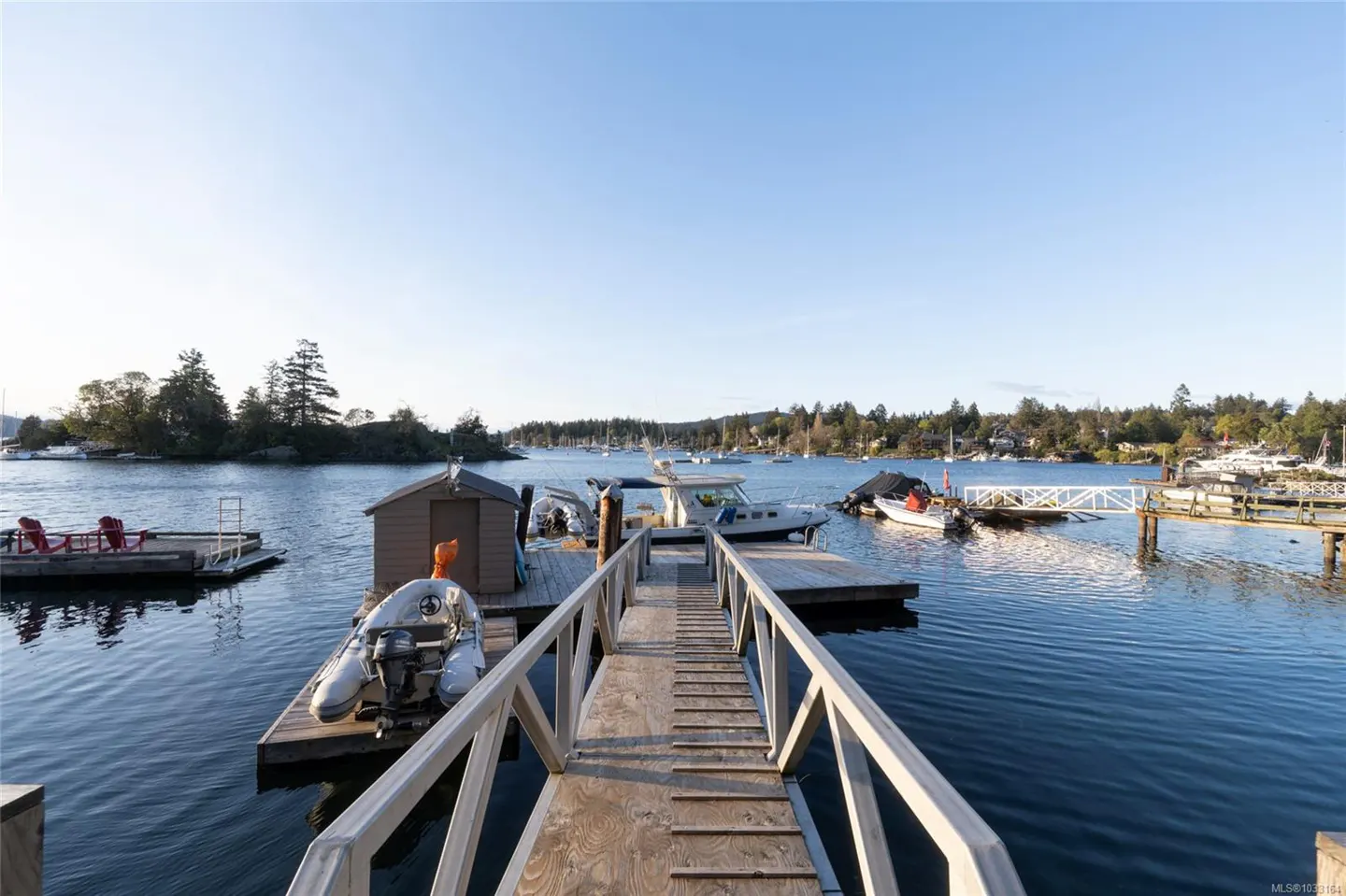 Waterfront view of a wooden dock with boats, a shed, and a calm blue bay under a clear sky.