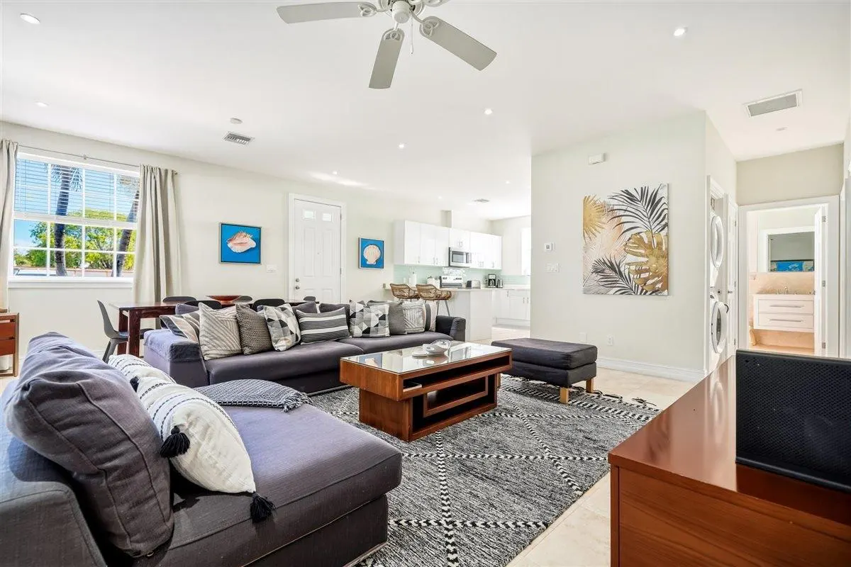 Bright, open-concept living room with a gray sectional sofa, wood coffee table, and a black and white patterned rug. White kitchen in the background.