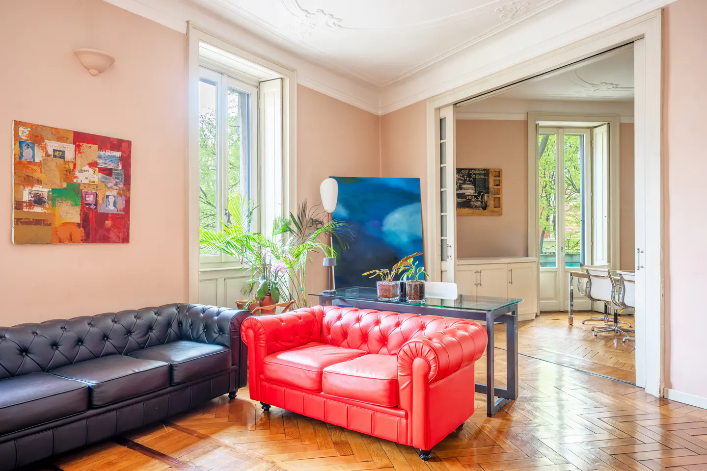 A living room with a black leather sofa, a red leather sofa, and a glass table with plants. A painting hangs on the wall.