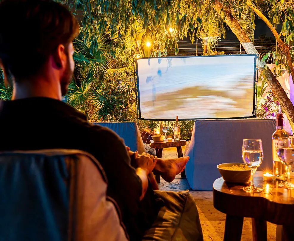 A man watches an outdoor movie on a screen surrounded by trees and lights. Drinks and snacks are on wooden tables.