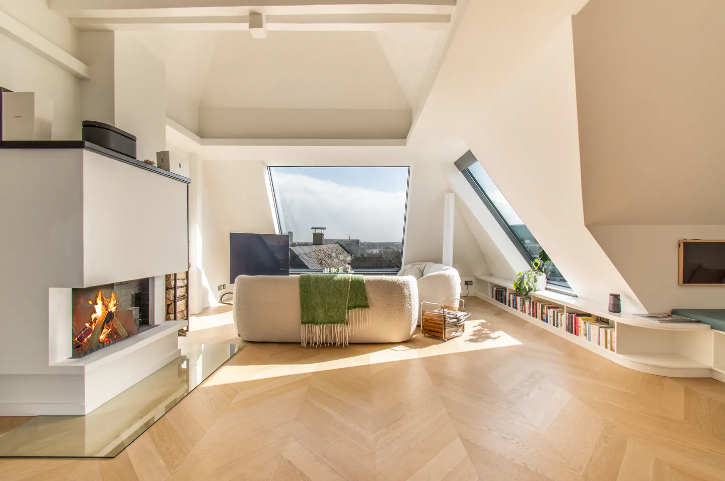 Bright, modern attic living room with a fireplace, herringbone wood floors, and large windows. A cream sofa has a green throw blanket.