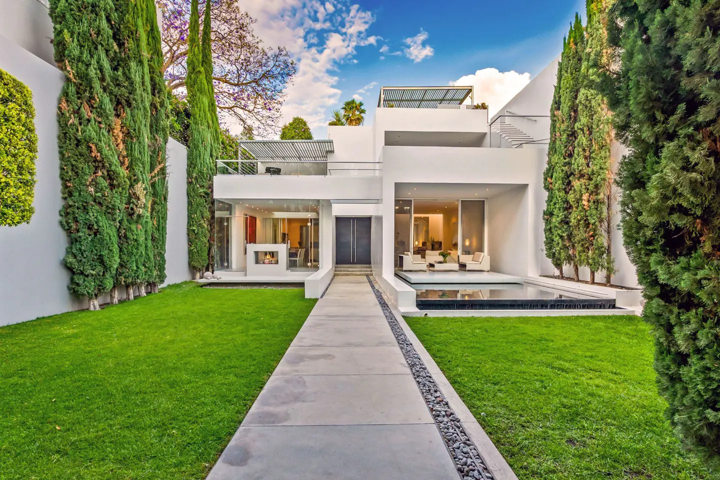 Modern white house with a walkway, green lawn, and tall trees under a blue sky with clouds.