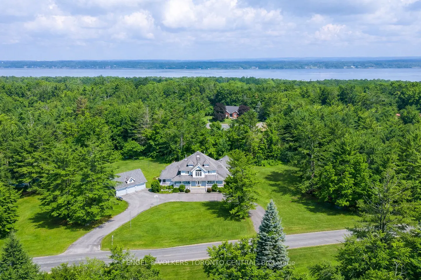 Aerial view of a large, light-blue house with a gray roof, surrounded by green trees and a lake in the background.