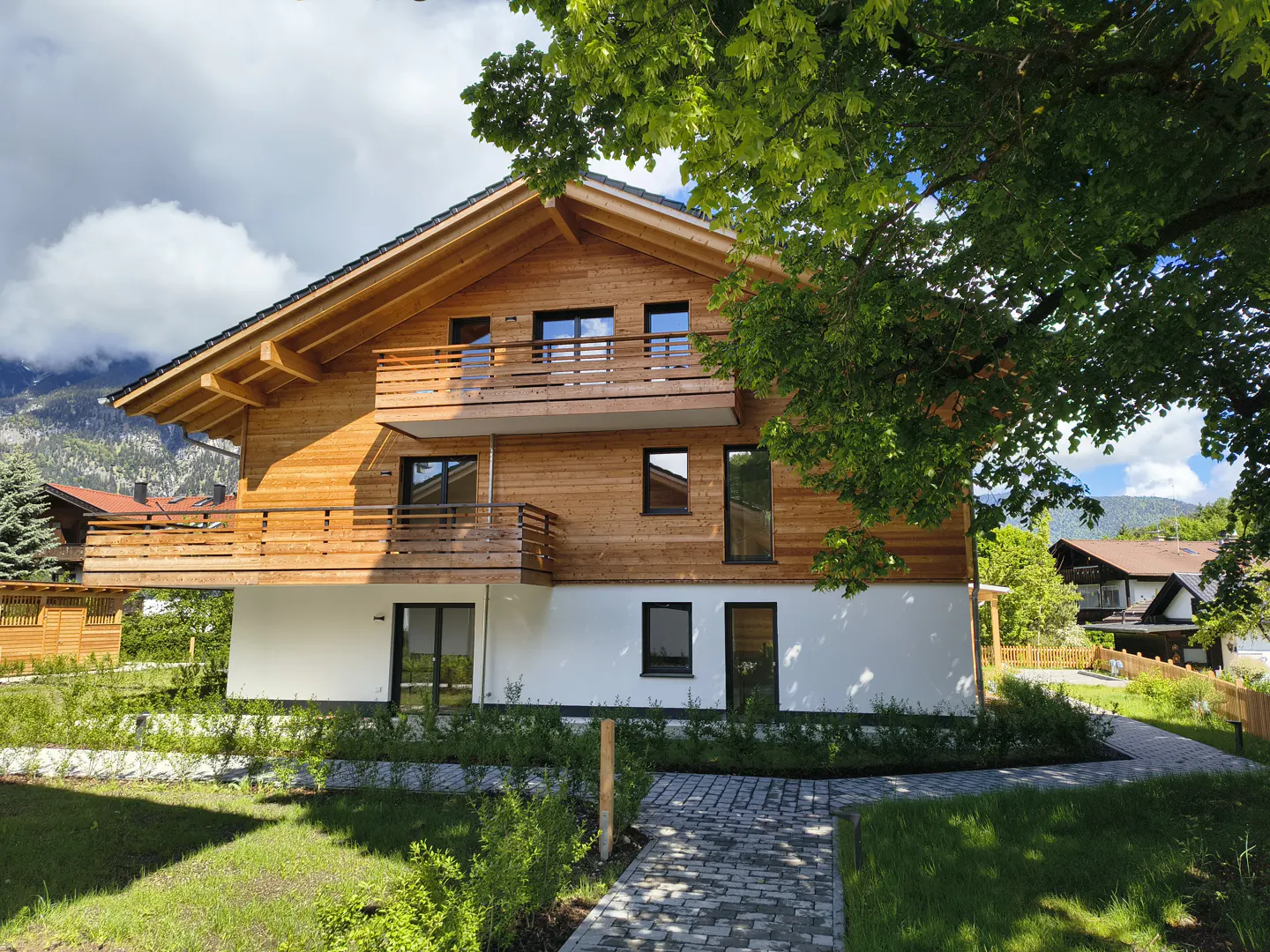 Two-story house with wood siding and white foundation, balcony, black framed windows, stone path, and green lawn.