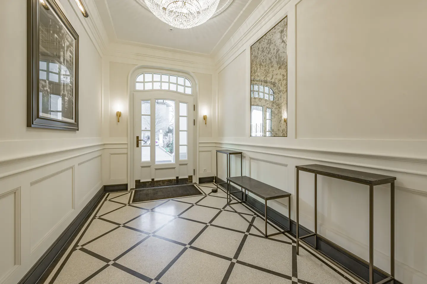 Elegant foyer with white walls, black and white checkered floors, and a crystal chandelier. A white door leads to the outside.