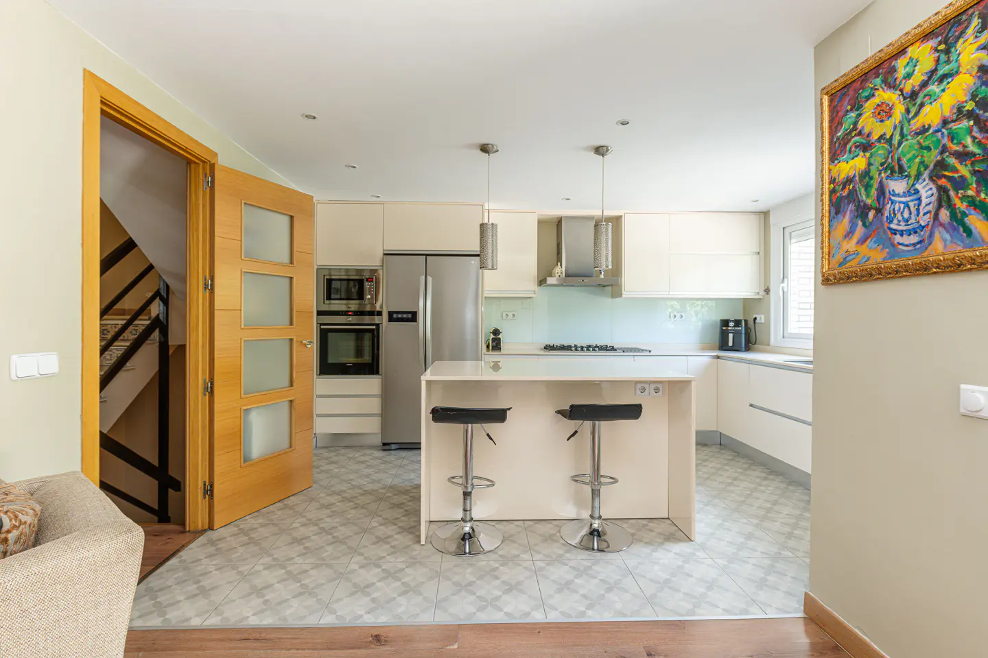 Bright kitchen with white cabinets, stainless steel appliances, and a center island with two black stools. A colorful sunflower painting hangs on the wall.