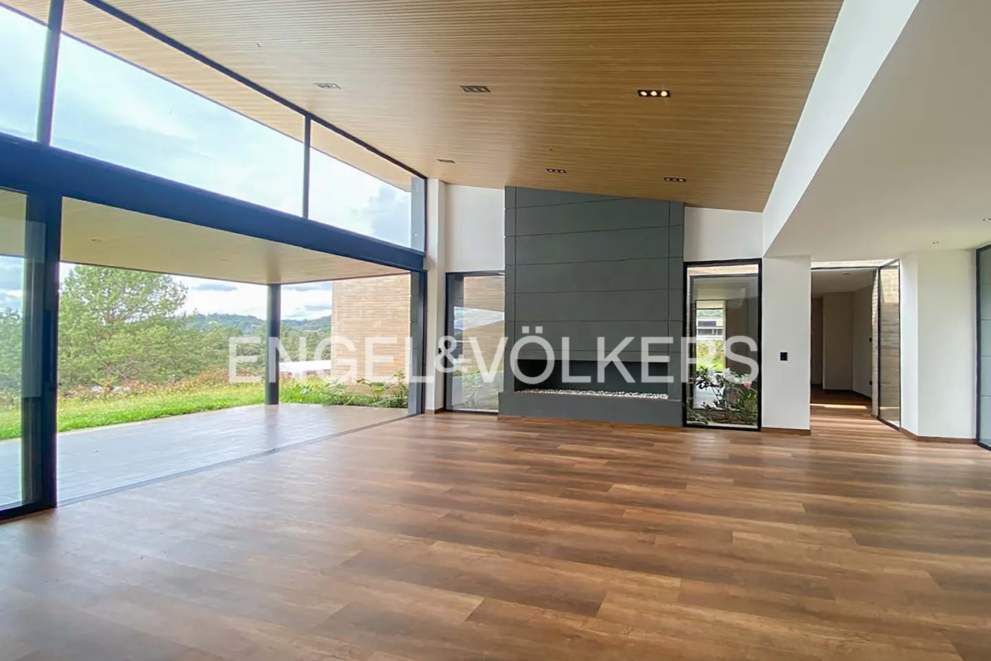 Modern living room with wood floors, a dark gray fireplace, and floor-to-ceiling windows overlooking a grassy landscape.