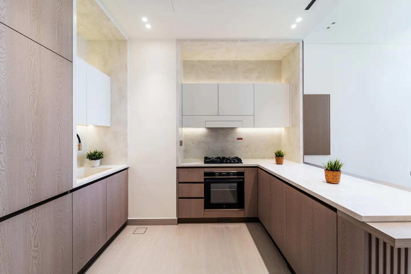 A modern kitchen with light wood cabinets, white countertops, and stainless steel appliances. Potted plants add a touch of greenery.