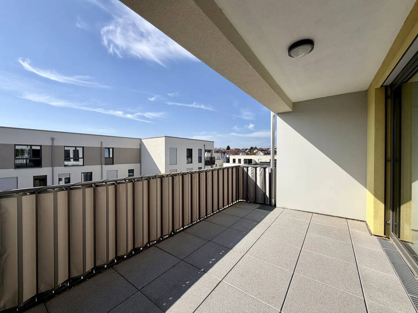 A sunny balcony with gray tiles, a privacy screen, and a view of nearby buildings under a blue sky.