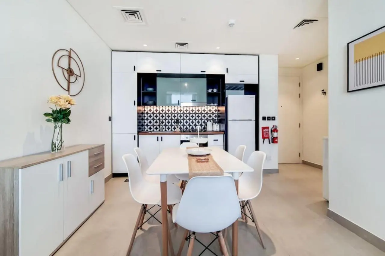 Modern dining area with a white table, four white chairs, and a kitchen featuring white cabinets and patterned backsplash.