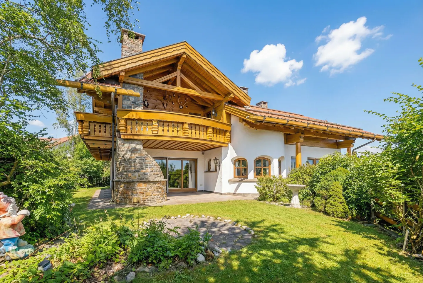 Exterior of a two-story chalet-style house with a stone chimney and wooden balcony, surrounded by green lawn and trees.