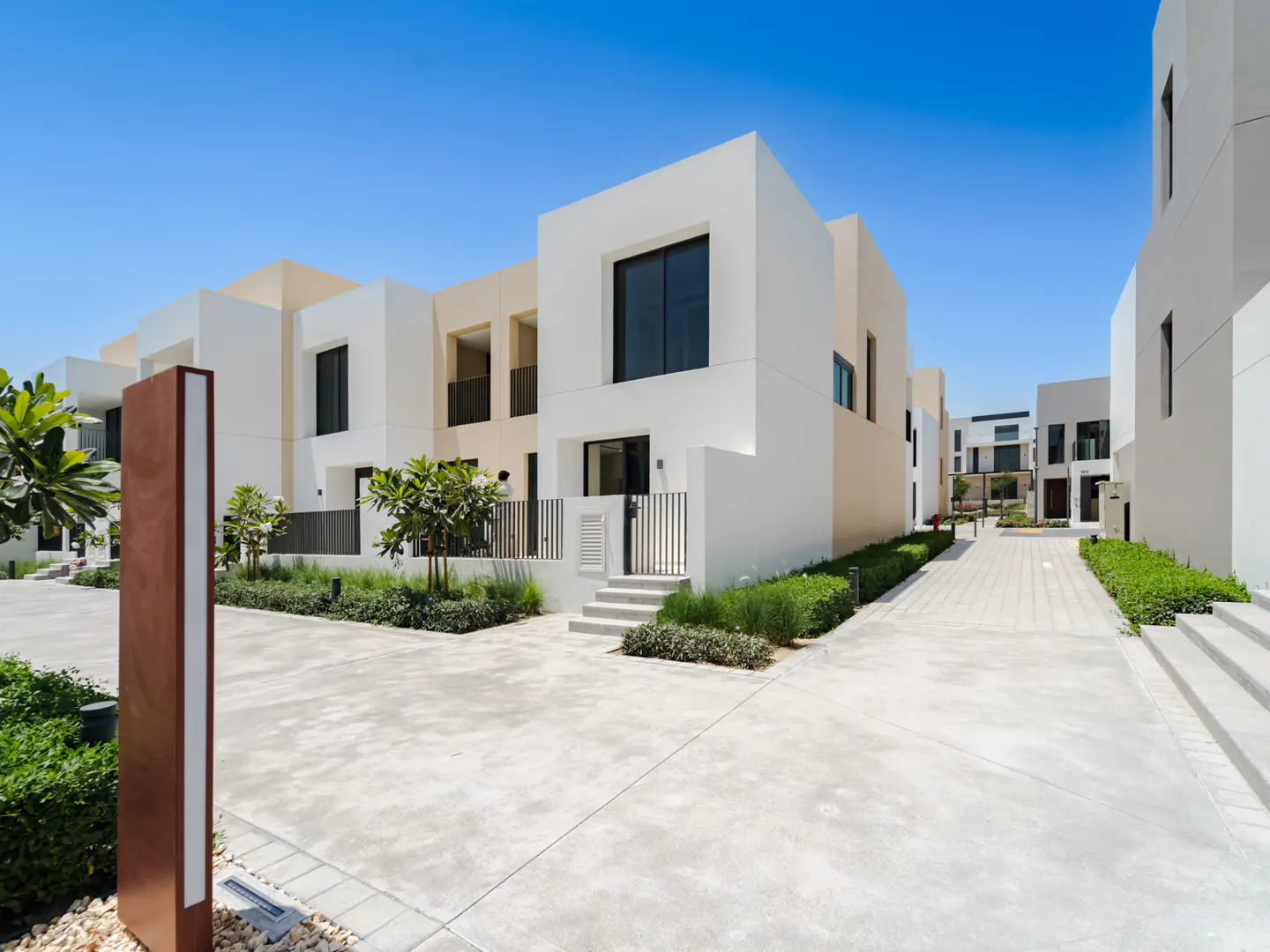 Modern white townhouses with black windows line a concrete walkway under a clear blue sky. Green landscaping adds a touch of nature.