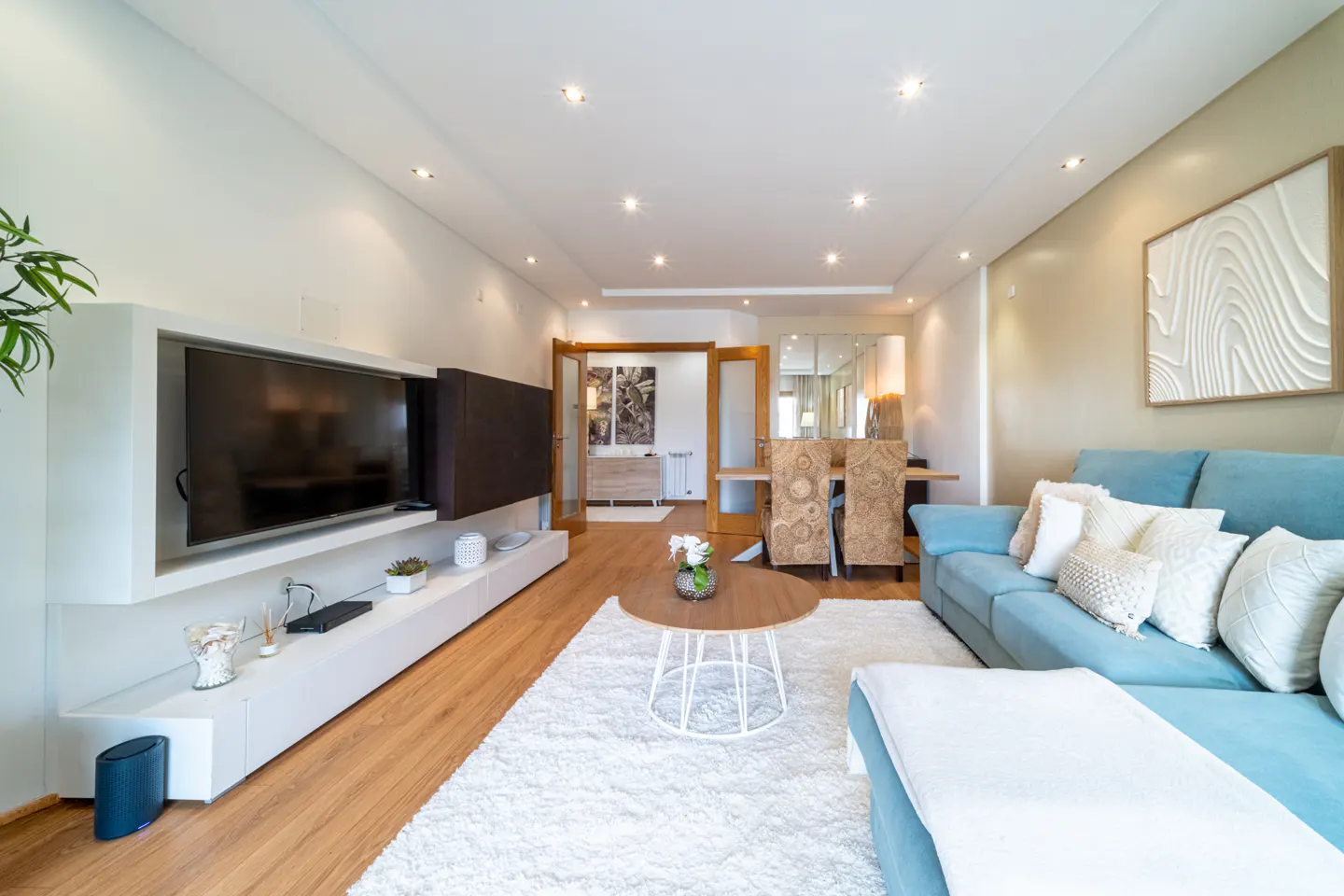 Bright living room with a blue sofa, white rug, and a modern TV stand. Dining area visible in the background.