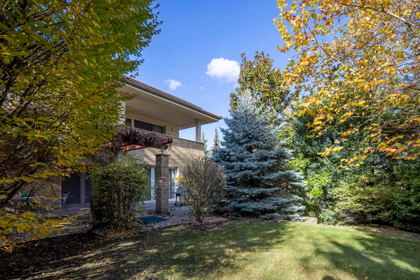 Two-story house with a balcony, surrounded by trees with green and yellow leaves, and a blue sky.