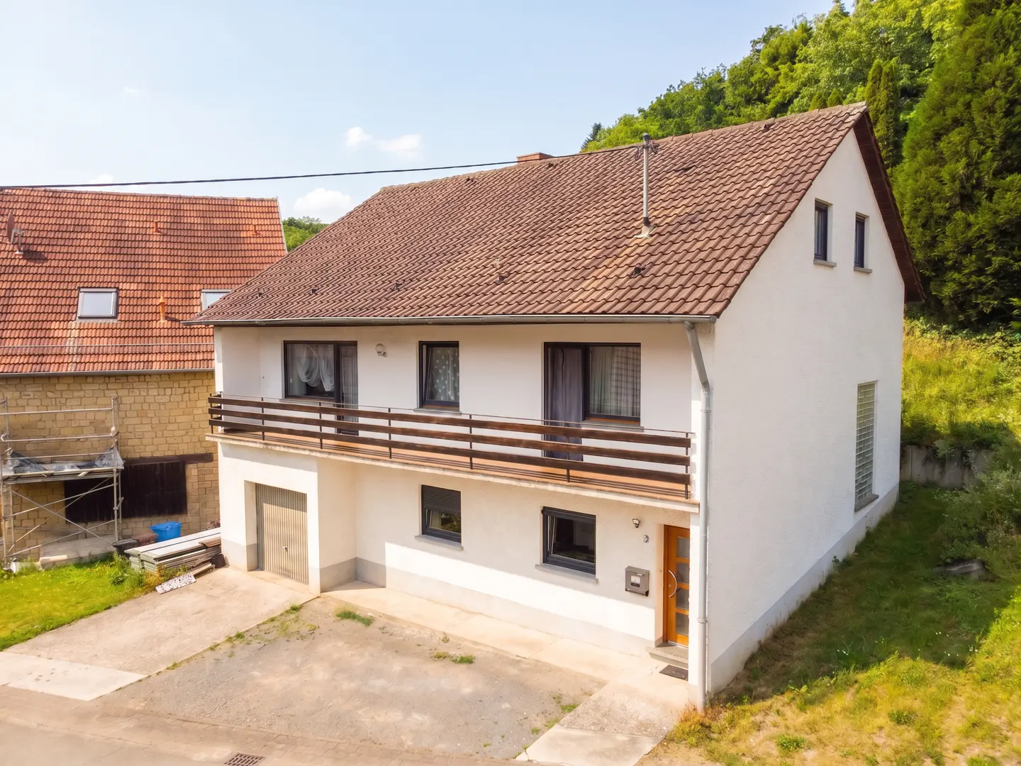 Two-story white house with a brown tiled roof, a wooden balcony, and an attached garage next to a stone building.