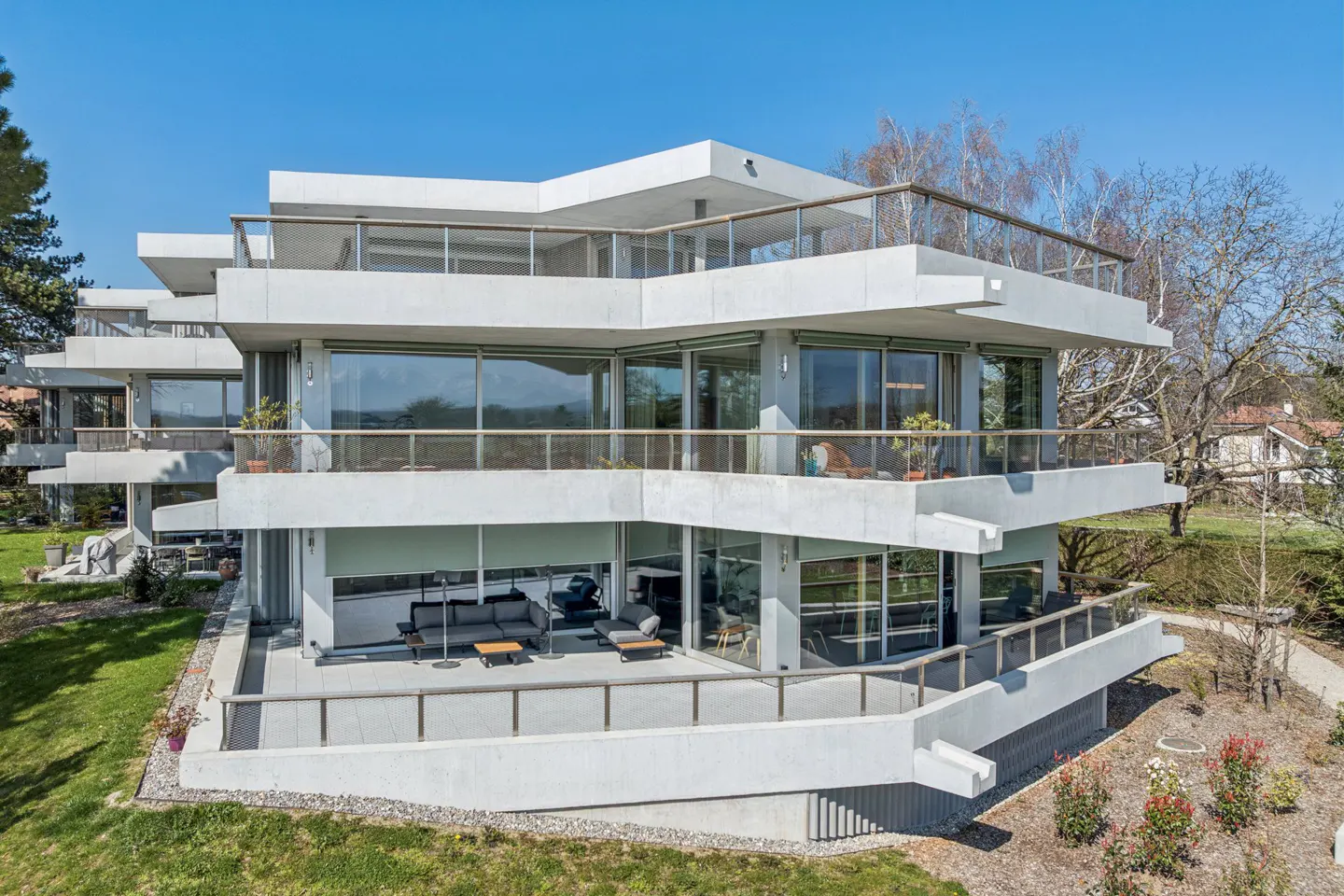Modern three-story concrete building with large windows and balconies. Outdoor furniture is visible on the balconies. Blue sky background.