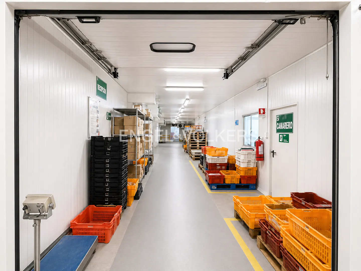 A long, brightly lit warehouse hallway with white walls and a gray floor. Crates and boxes line the walls.