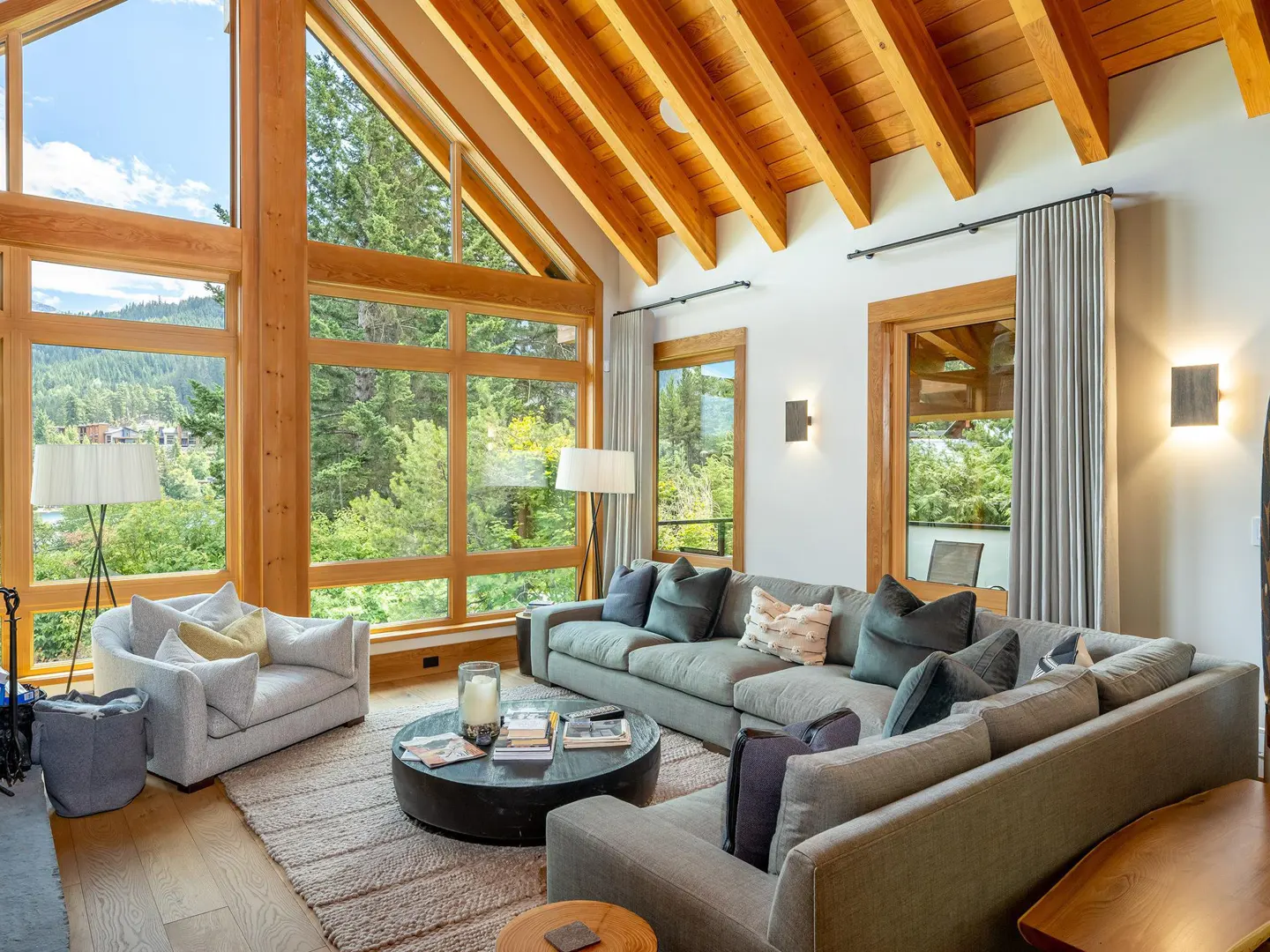 Bright living room with wood-framed windows overlooking a forest. A large gray sectional sofa and armchair sit on a textured rug. The ceiling has exposed wood beams.