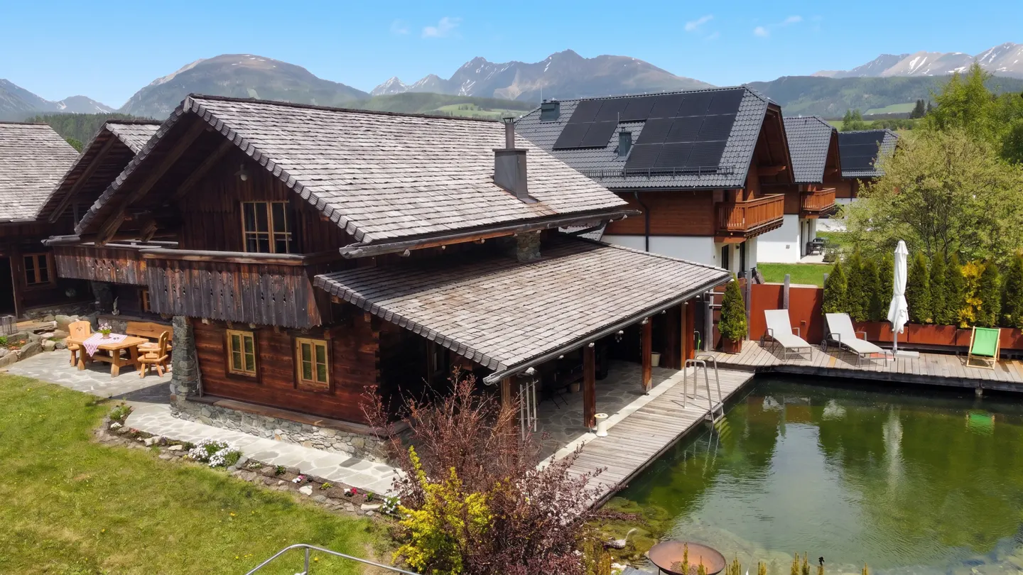 Rustic wooden chalet with a gray shingle roof, next to a pond with a wooden deck and lounge chairs, set against a mountain backdrop.