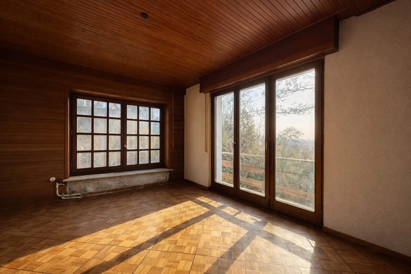 Empty room with wood paneled walls, ceiling, and parquet floor. Sunlight streams in from a window and glass doors.