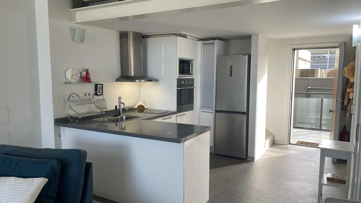 A modern kitchen with white cabinets, stainless steel appliances, and a gray countertop island. A doorway leads to an outdoor balcony.