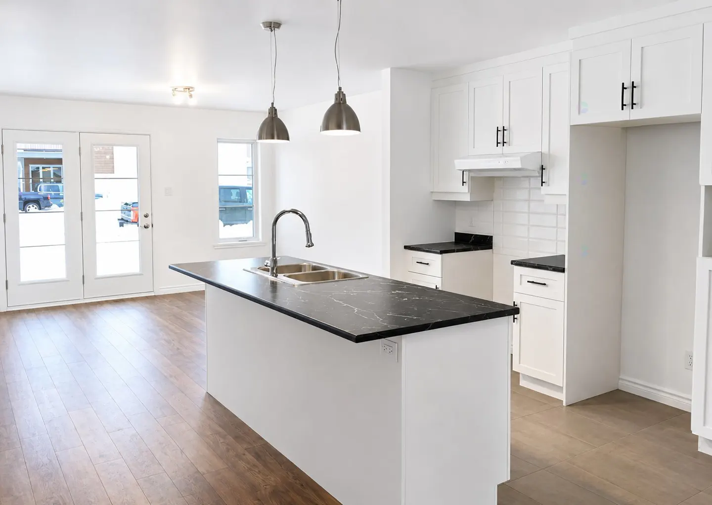 Bright, modern kitchen with white cabinets, black countertops, and stainless steel fixtures. Island with sink and pendant lights. Wood floors and white walls.