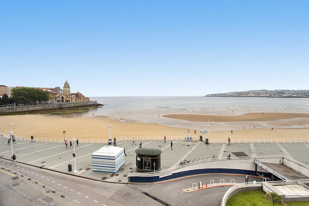 High-angle view of a sandy beach promenade with pedestrians, a striped beach hut, and a coastal town under a clear blue sky.