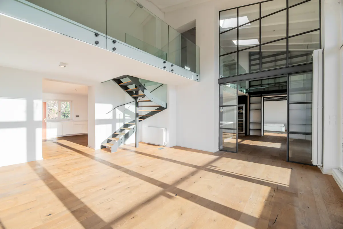 Bright, modern loft interior with hardwood floors, spiral staircase, glass railing, and black-framed glass wall. Sunlight streams through windows.