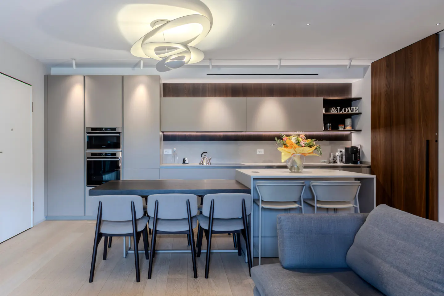 A modern kitchen with gray cabinets, a dark countertop island with four chairs, and a white island with bar stools. A gray sofa is in the foreground.