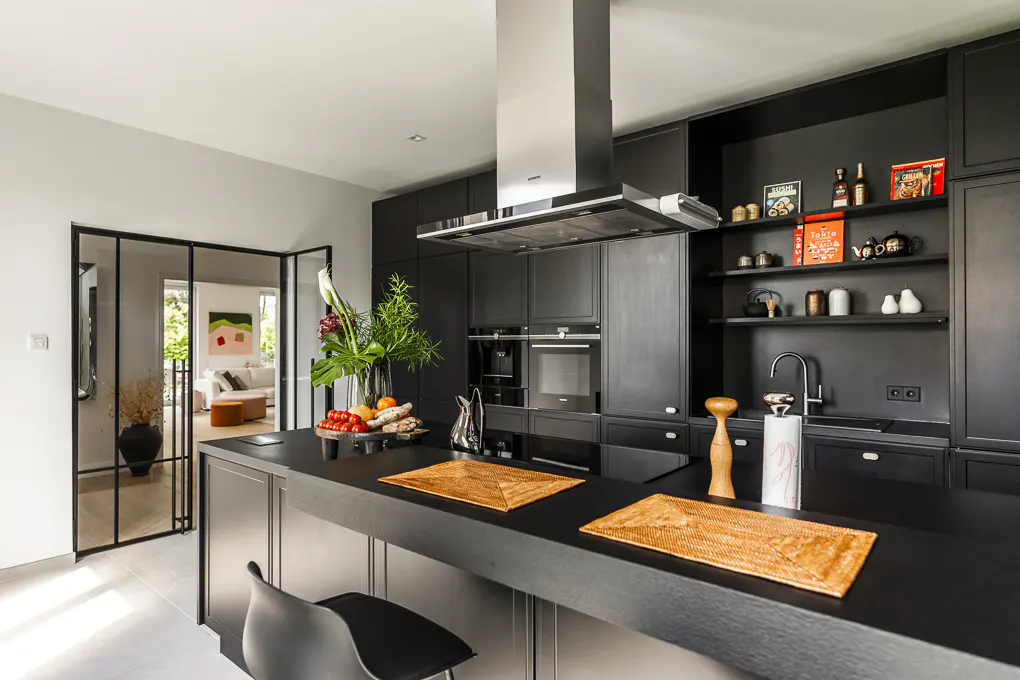 A modern kitchen with black cabinets, stainless steel hood, and black island. A glass door leads to a living room with a white sofa.
