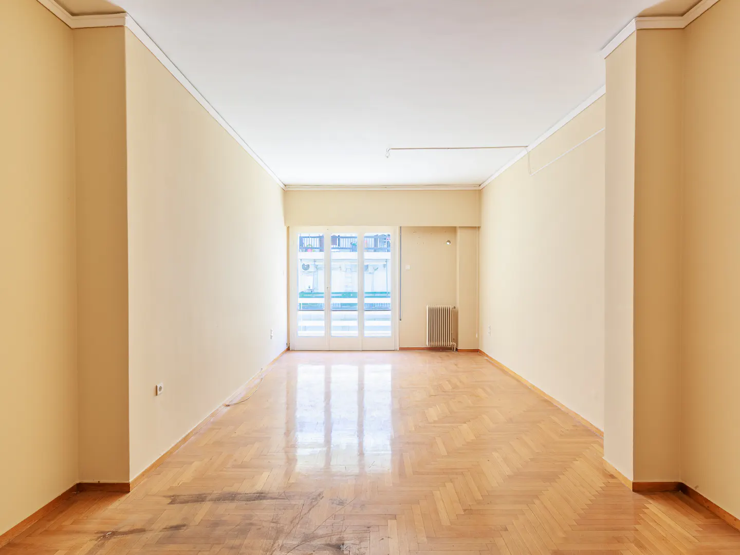 Empty room with parquet floor, cream walls, and white trim. Balcony doors at the end of the room let in natural light.