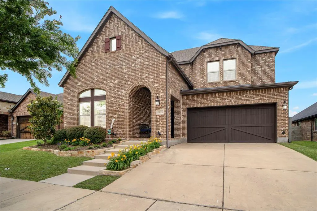 Two-story brick house with an arched entryway, a dark brown garage door, and a well-manicured lawn with yellow flowers.
