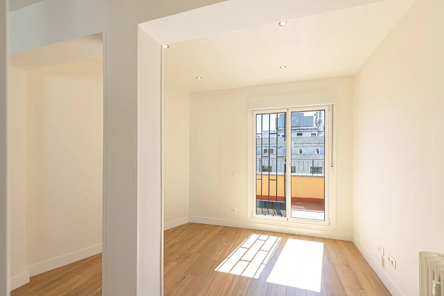 Bright, empty room with wood floors and white walls. A window with bars looks out onto a city scene. Sunlight streams in.