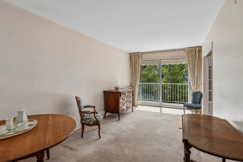 Bright living room with beige carpet, antique wooden furniture, patterned chairs, and a balcony view of green trees.