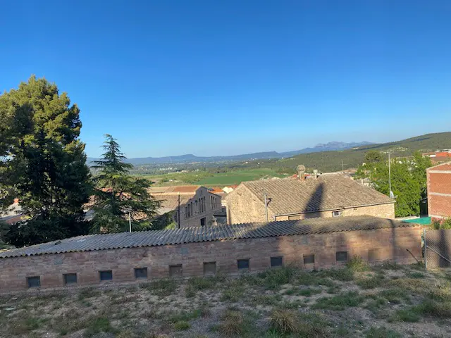 View of a rural landscape with a brick wall in the foreground, buildings with tiled roofs, green fields, trees, and mountains under a clear blue sky.