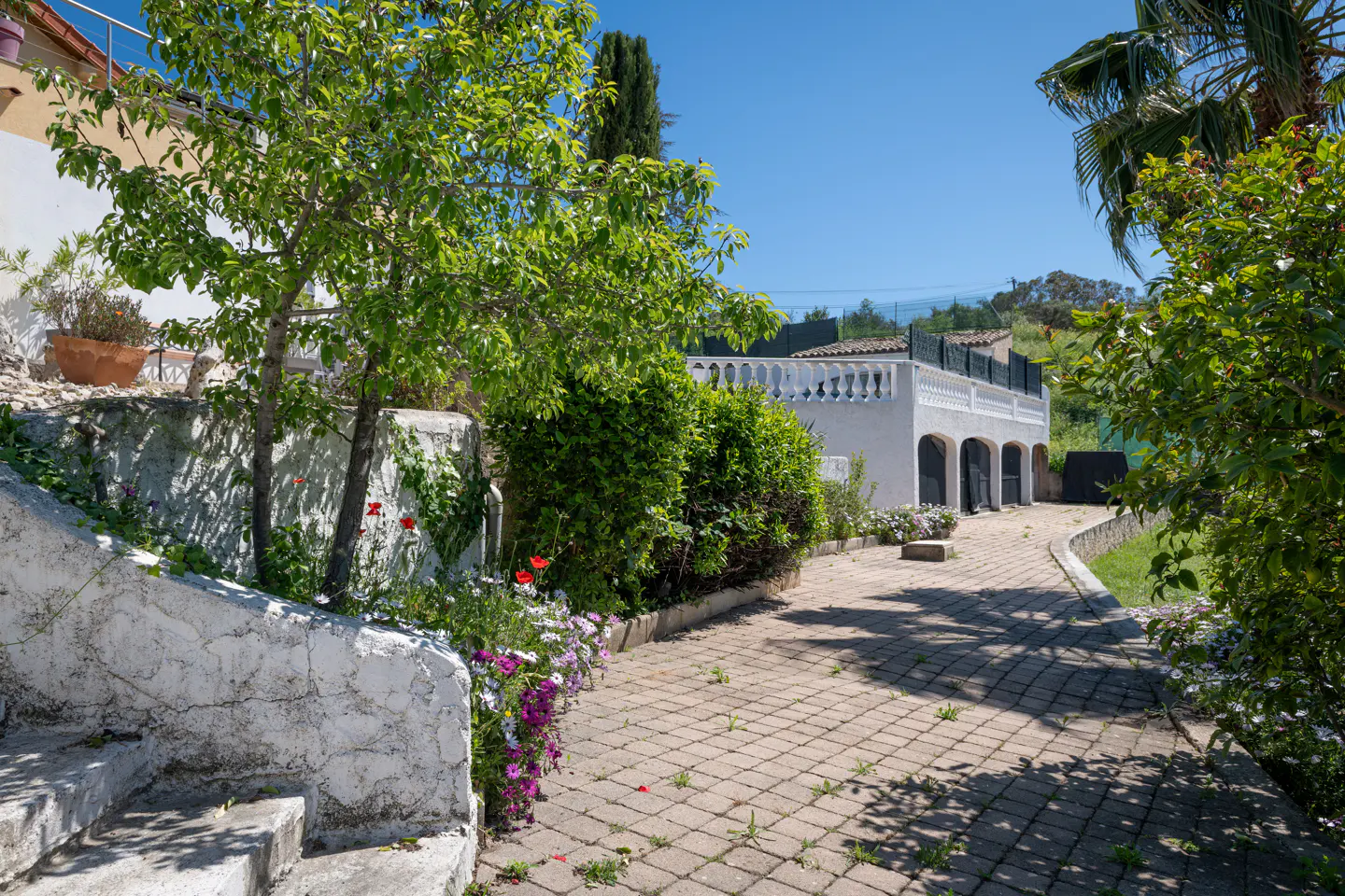 Exterior view of a white building with arched doorways and a brick-paved driveway, surrounded by lush greenery and flowers.