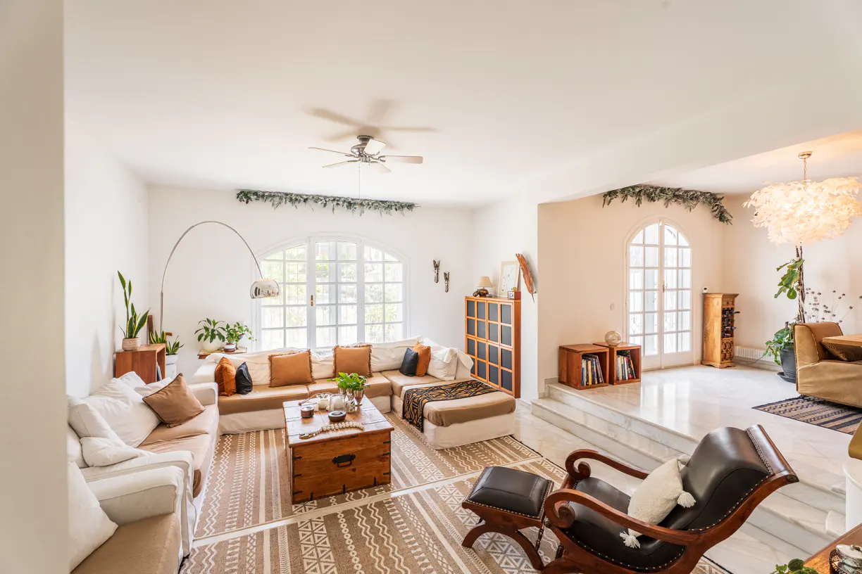 Bright living room with white walls, arched windows, and a large sectional sofa. A wooden chest serves as a coffee table on a patterned rug.