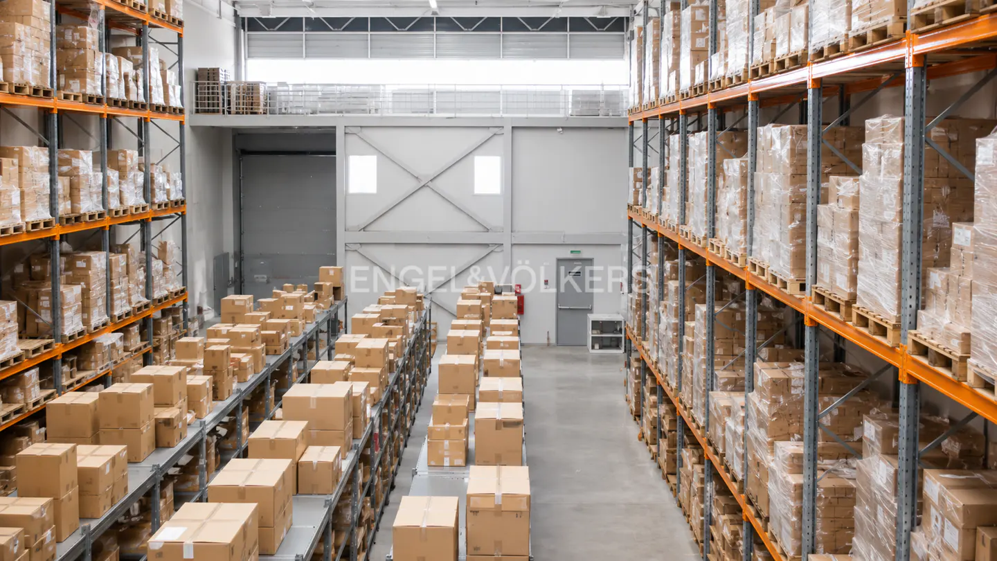 Warehouse interior with rows of tall shelves filled with cardboard boxes. Gray concrete floor and walls. Engel & Völkers logo visible.