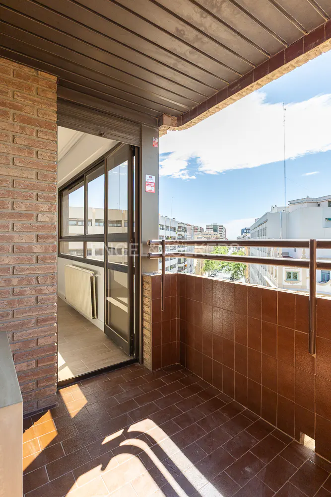 A balcony with brown tile flooring and a tiled wall overlooks a city skyline under a blue sky.